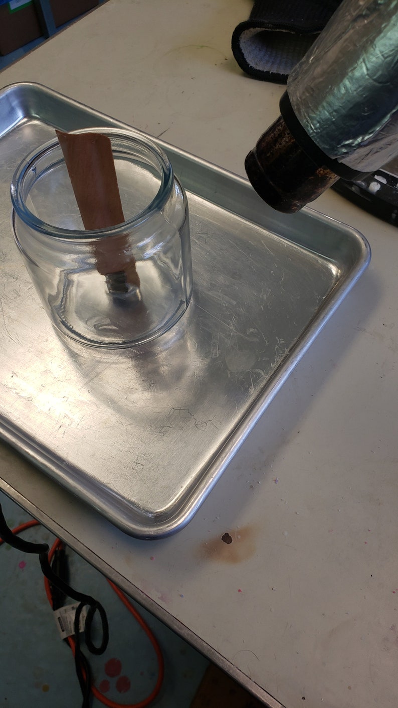May include: A heat gun is being used to heat a glass jar with a brown paper strip inside. The jar is sitting on a silver baking sheet.