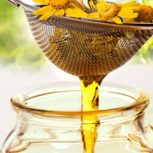 May include: A clear glass jar filled with golden liquid.  A strainer with yellow flowers is pouring the liquid into the jar.