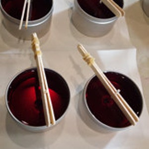May include: Four red candles in silver tins with wooden wicks secured with rubber bands. The candles are on a white surface.