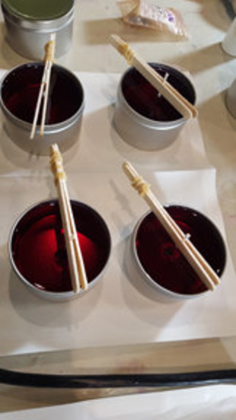 May include: Four red candles in silver tins with wooden wicks secured with rubber bands. The candles are on a white surface.