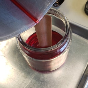 May include: A glass jar filled with a red liquid, a wooden stick is in the jar, and a metal container is pouring the liquid into the jar.
