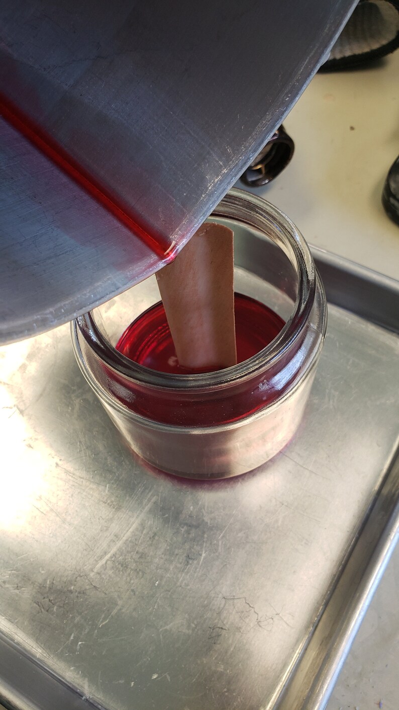 May include: A glass jar filled with a red liquid, a wooden stick is in the jar, and a metal container is pouring the liquid into the jar.