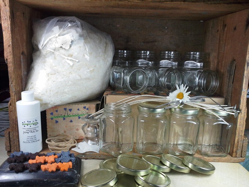 Puede incluir: Una caja de madera llena de suministros para hacer velas. Los suministros incluyen una bolsa de copos de cera blanca, una botella de aceite de fragancia, frascos de vidrio, tapas, mechas y gomas el&aacute;sticas.