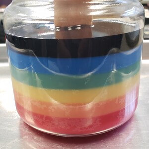 May include: A glass jar filled with layers of colorful wax, creating a rainbow effect. The wax is red, orange, yellow, green, blue, and black.