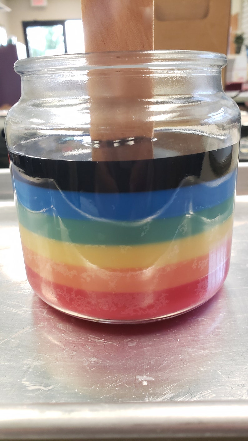 May include: A glass jar filled with layers of colorful wax, creating a rainbow effect. The wax is red, orange, yellow, green, blue, and black.
