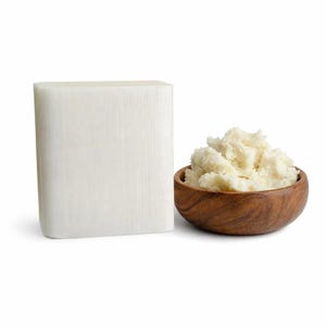 May include: A rectangular block of solid white shea butter positioned next to a small, round, dark brown wooden bowl filled with a mound of soft, creamy, off-white shea butter with a textured, uneven surface and a natural appearance.