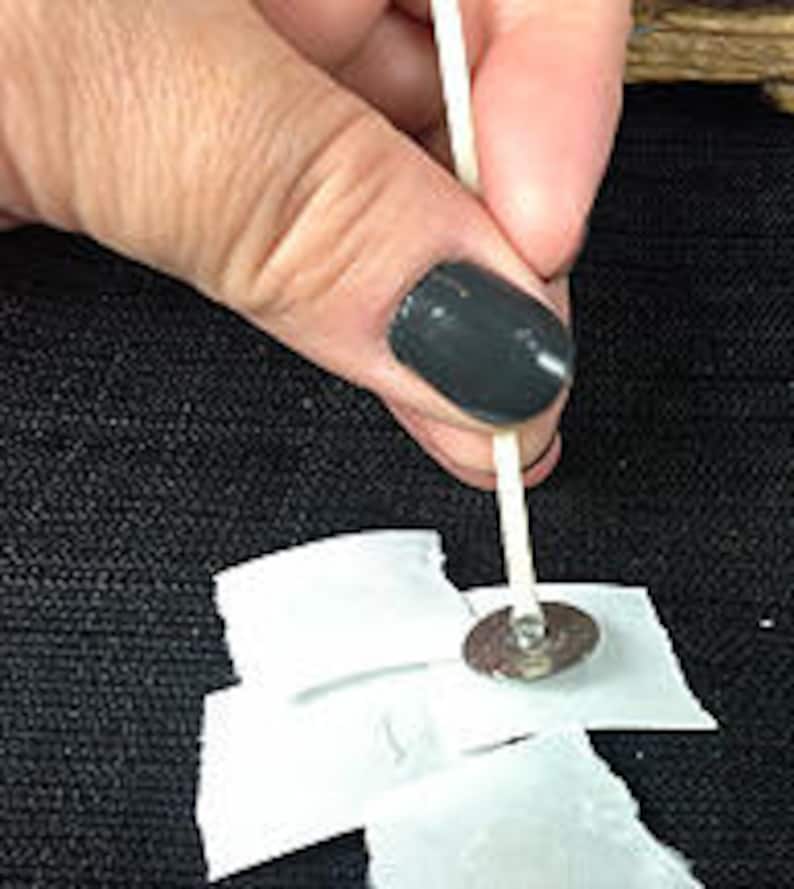 May include: A white cotton wick is threaded through a silver metal candle base, secured with a white adhesive sticker on a black fabric surface.