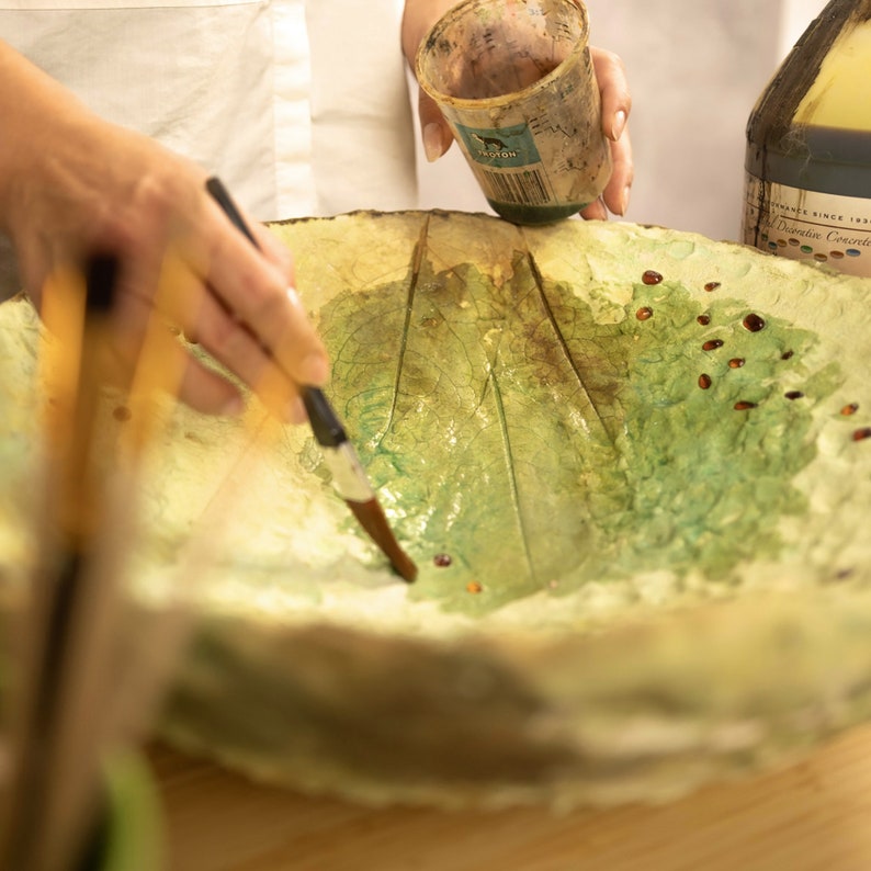 May include: A person is shown painting a large, leaf-shaped bowl with a paintbrush. The bowl is primarily green and yellow, with visible leaf vein details. Small, reddish-brown droplets are scattered across the surface. A cup and bottle are also visible.