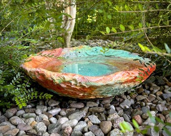 May include: A colorful, handcrafted bird bath with a textured surface. The bird bath features a shallow basin filled with water, surrounded by a rim painted in shades of orange, green, and pink. It rests on a bed of small stones, surrounded by greenery.