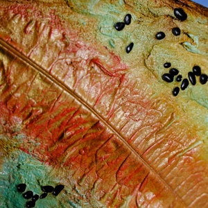May include: Close-up of a textured, abstract artwork featuring a leaf-like design. The artwork uses a palette of warm colors, including gold, red, and orange, with accents of green and black. The surface has a rough, organic appearance.