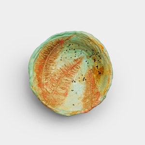 May include: A handmade, irregularly shaped bowl with a textured surface. The bowl features a blend of colors, including green, gold, and orange, with small black speckles. The interior design resembles a stylized leaf pattern.
