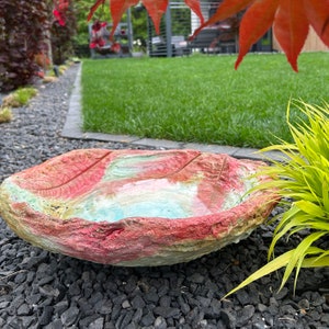 May include: A leaf-shaped bird bath made of concrete, featuring a textured surface with red, pink, and green hues. The bird bath is filled with water and rests on a bed of black gravel, with a green lawn in the background.
