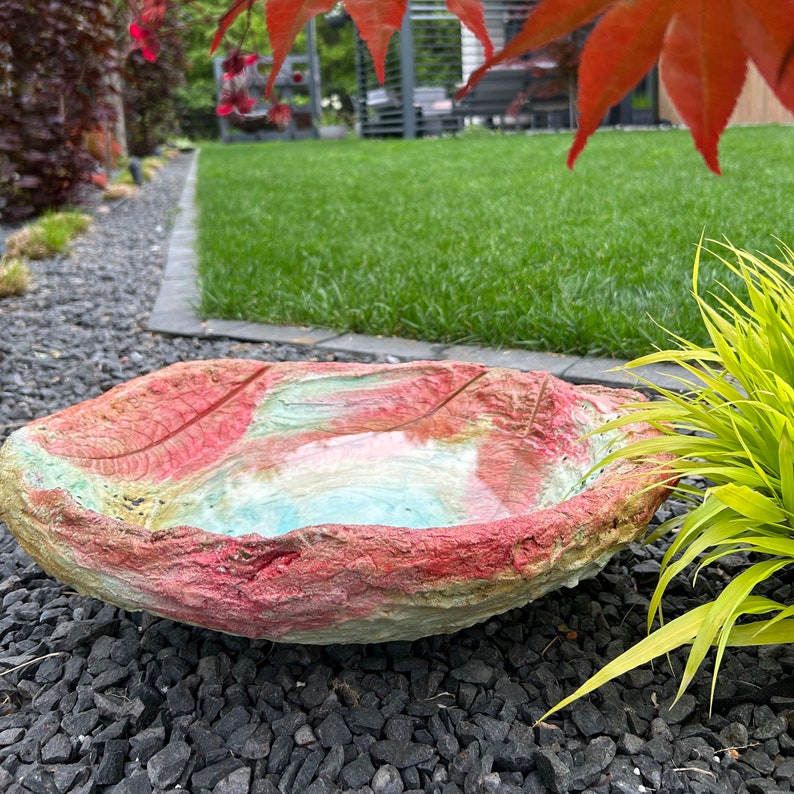 May include: A leaf-shaped bird bath made of concrete, featuring a textured surface with red, pink, and green hues. The bird bath is filled with water and rests on a bed of black gravel, with a green lawn in the background.