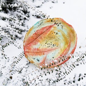 May include: A round, hand-painted decorative bowl with a leaf-like design in shades of red, orange, yellow, and blue. The bowl is set in a snowy outdoor scene, surrounded by green foliage. The bowl's diameter is approximately 12 inches.