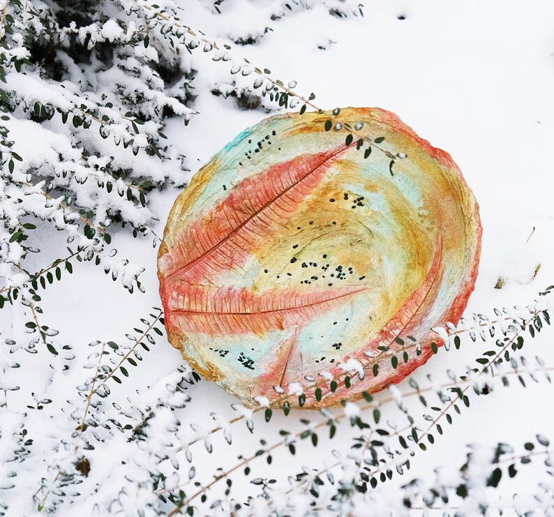 May include: A round, hand-painted decorative bowl with a leaf-like design in shades of red, orange, yellow, and blue. The bowl is set in a snowy outdoor scene, surrounded by green foliage. The bowl's diameter is approximately 12 inches.