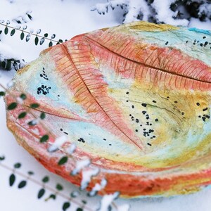 May include: A colorful, leaf-shaped decorative bowl with a textured surface. The bowl features a painted design with shades of red, yellow, blue, and green. Small black accents are scattered across the surface. The bowl is set against a snowy background.