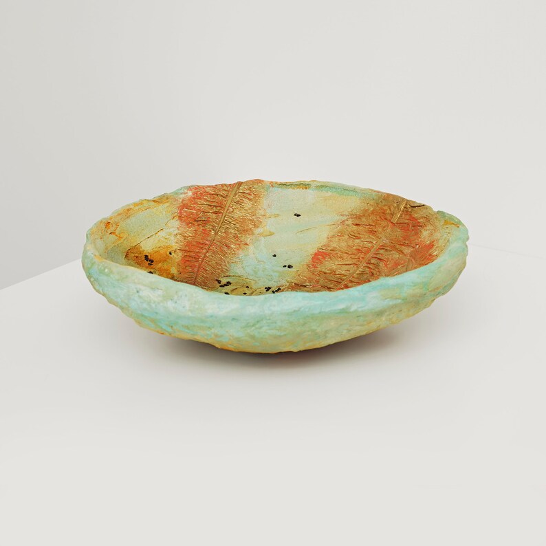 May include: A handmade, oval-shaped ceramic bowl with a textured surface. The bowl features a light blue rim and a center with orange and gold leaf patterns. Small black specks are scattered throughout the interior.