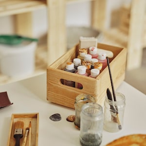 May include: A wooden crate filled with paint bottles, brushes, and jars. The paint bottles have white and gold lids, and the brushes are in a small wooden tray. Seashells are scattered on the white surface.