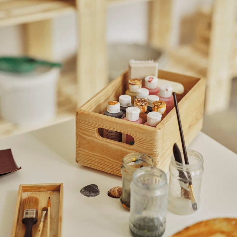 May include: A wooden crate filled with paint bottles, brushes, and jars. The paint bottles have white and gold lids, and the brushes are in a small wooden tray. Seashells are scattered on the white surface.