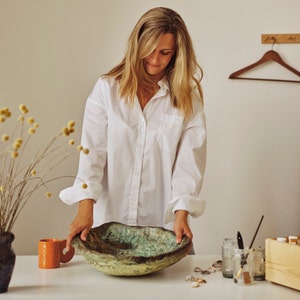 May include: A large, handcrafted ceramic bowl with a textured surface and a blend of earthy tones, including brown, green, and gold. The bowl is held by a person wearing a white shirt. A small orange mug and dried flowers are also visible.