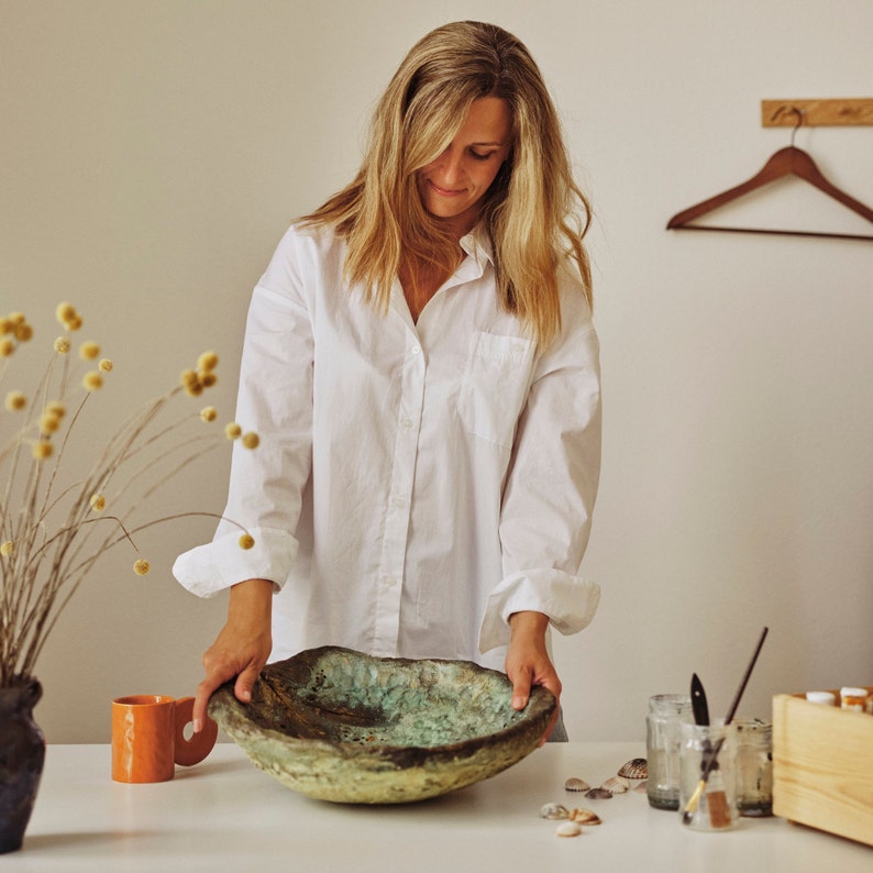 May include: A large, handcrafted ceramic bowl with a textured surface and a blend of earthy tones, including brown, green, and gold. The bowl is held by a person wearing a white shirt. A small orange mug and dried flowers are also visible.