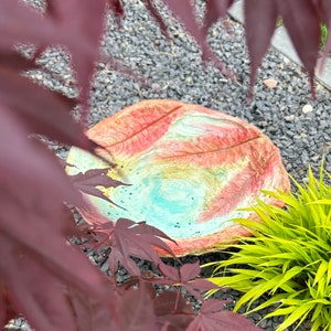 May include: A colorful, round bird bath with a textured surface. The bird bath features a design with red, orange, yellow, and turquoise hues. The bird bath is surrounded by dark gray gravel, green grass, and red leaves.
