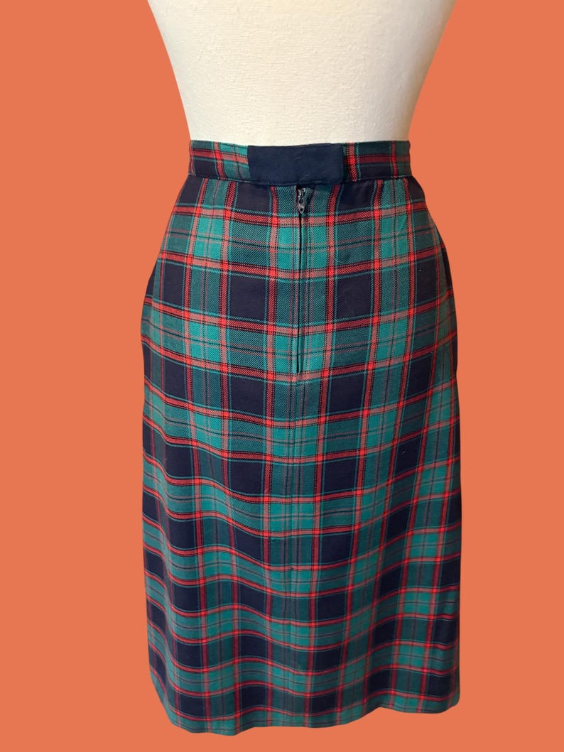 May include: A plaid pencil skirt in a classic tartan pattern. The skirt features a navy blue, green, and red color scheme. It has a zipper closure at the back and a navy blue waistband. The skirt is displayed on a mannequin.