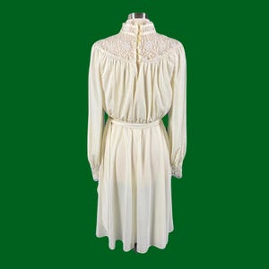 Pode incluir: Um vestido branco de manga comprida com colarinho e punhos de renda. O vestido tem um corpete justo e uma saia fluida.