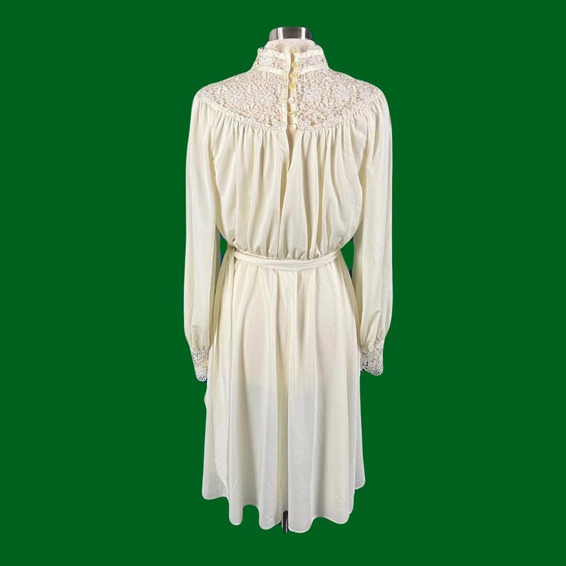Pode incluir: Um vestido branco de manga comprida com colarinho e punhos de renda. O vestido tem um corpete justo e uma saia fluida.