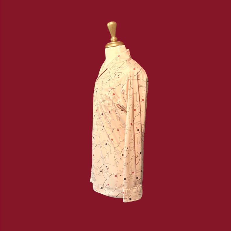 May include: A light peach-colored long-sleeve shirt with a geometric pattern of black lines and red and black squares. The shirt is buttoned and has a collar and a breast pocket. The shirt is displayed on a mannequin.