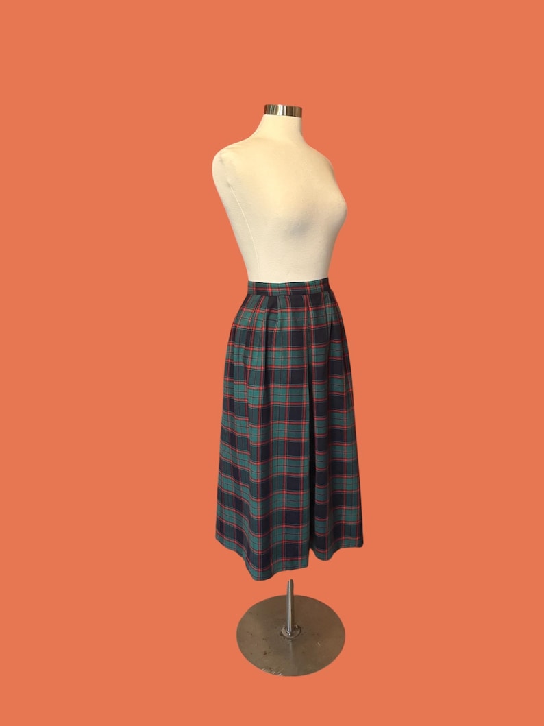 May include: A vintage plaid skirt in green, navy, and red, displayed on a mannequin. The skirt has a pleated design and falls to a mid-calf length. The fabric features a classic tartan pattern, showcasing a timeless style.