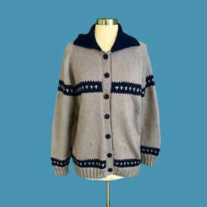 Op de afbeelding: Een grijze gebreide cardigan met een blauwe kraag en blauwe en witte gepatroneerde banden rond de manchetten en taille.