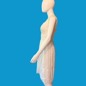 May include: A cream-colored sleeveless dress with delicate lace detailing. The dress features a fitted bodice and a flowing skirt, with lace trim at the hem. The dress is displayed on a mannequin against a blue background.