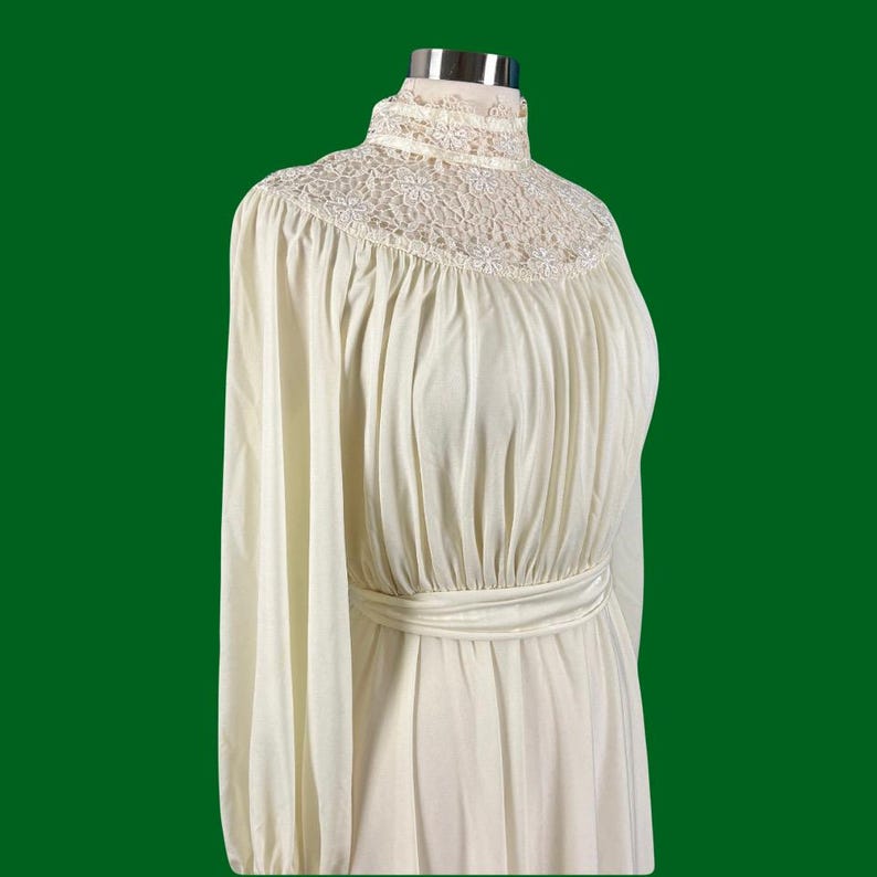 Pode incluir: Um vestido branco de manga comprida com gola alta e acabamento em renda. O vestido tem um corpete drapeado e uma cintura ajustada.