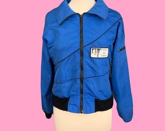 May include: A bright blue zip-up jacket featuring a black front zipper, black ribbed hem and cuffs, and a white rectangular patch on the chest with the text "Catskill Ski School". The jacket has black piping details across the front and long sleeves.