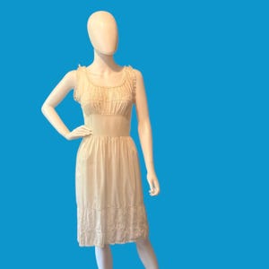 May include: Cream-colored vintage slip dress on a mannequin. The dress features a square neckline with lace trim, a fitted bodice, and a flowing skirt. The hemline is also trimmed with lace. The dress is displayed against a bright blue background.