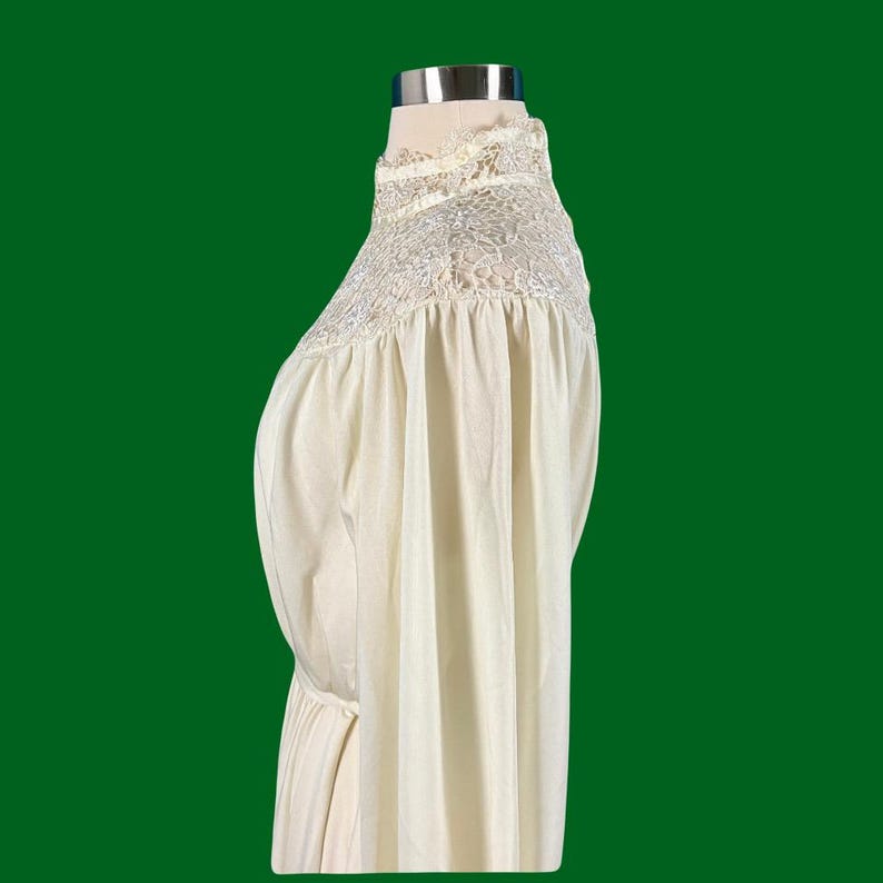 Pode incluir: Um vestido branco de manga comprida com gola alta e detalhes em renda. O vestido tem uma silhueta fluida e uma cintura franzida.