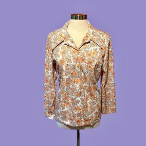 May include: A vintage floral print blouse with long sleeves and a collared neckline. The blouse features a white background with a repeating pattern of orange and yellow flowers. The buttons are orange, and the blouse is displayed on a mannequin.