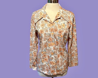 Puede incluir: Una blusa vintage con estampado floral, mangas largas y cuello. La blusa presenta un fondo blanco con un patrón repetido de flores naranjas y amarillas. Los botones son naranjas y la blusa se exhibe en un maniquí.