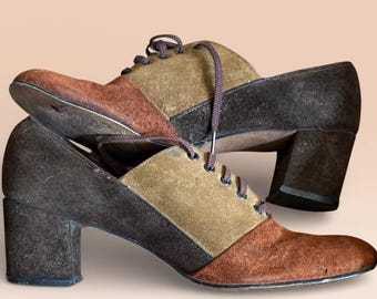 Puede incluir: Un par de zapatos vintage de ante de dos tonos con tacones de bloque. Los zapatos presentan paneles marrones, beige y marrón oscuro, con cordones a juego. Los tacones miden aproximadamente 6,35 cm de alto.