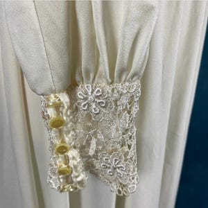 Pode incluir: Um vestido branco de manga comprida com uma borda de renda nos punhos. Os punhos t&ecirc;m quatro bot&otilde;es amarelos.