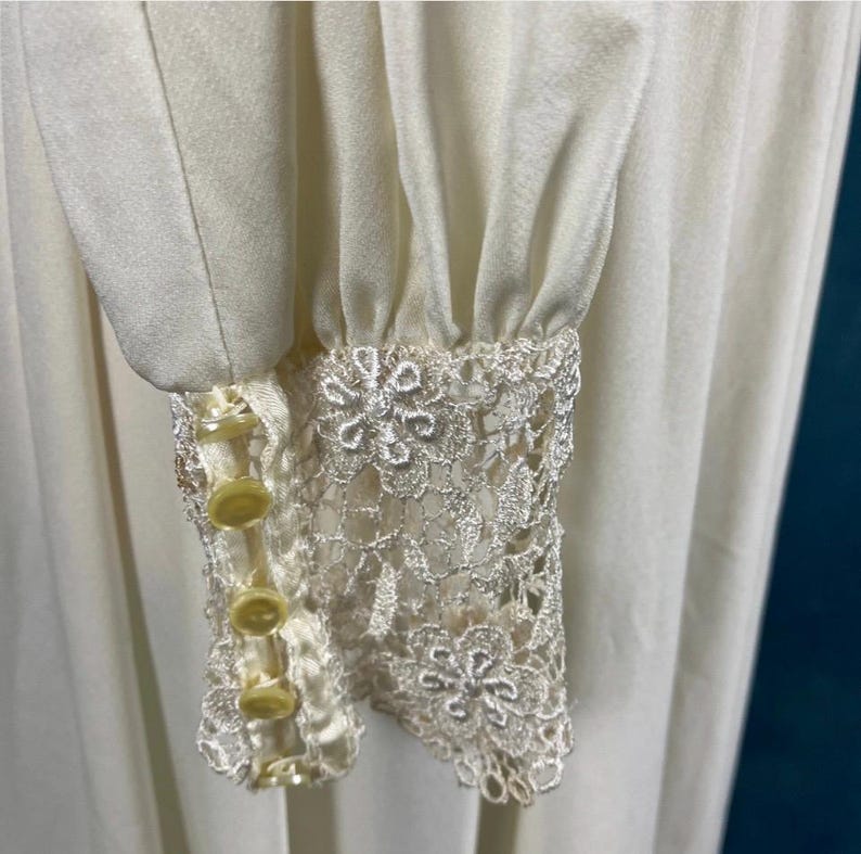 Pode incluir: Um vestido branco de manga comprida com uma borda de renda nos punhos. Os punhos t&ecirc;m quatro bot&otilde;es amarelos.