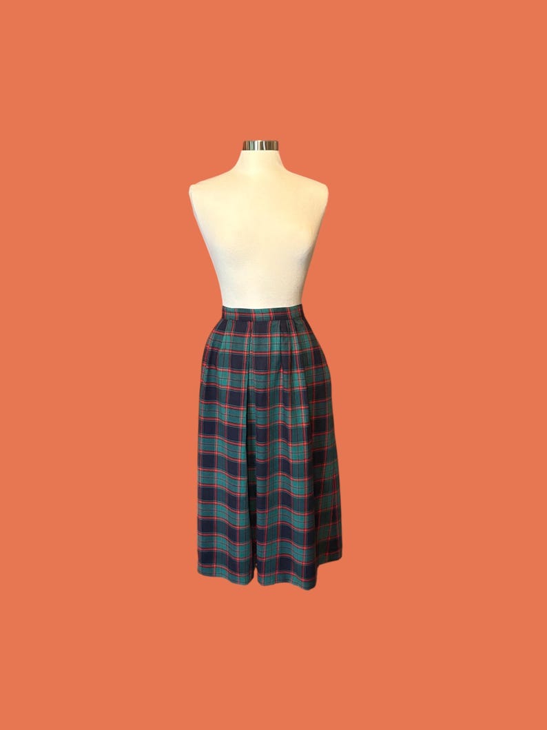May include: A vintage plaid skirt in shades of green, navy, and red, displayed on a mannequin. The skirt features a classic pleated design and a high waist. The fabric has a traditional tartan pattern, perfect for a retro or preppy look.
