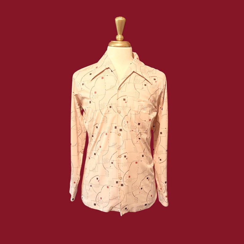 May include: A light peach-colored button-down shirt with a geometric pattern of black, brown, and red shapes. The long-sleeved shirt has a collared neckline and a single chest pocket. The shirt is displayed on a mannequin against a dark red background.