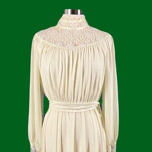 Pode incluir: Um vestido branco marfim de manga comprida com gola alta e acabamento em renda. O vestido tem um corpete justo e uma saia fluida.