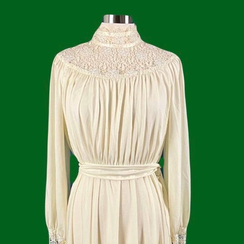 Pode incluir: Um vestido branco marfim de manga comprida com gola alta e acabamento em renda. O vestido tem um corpete justo e uma saia fluida.
