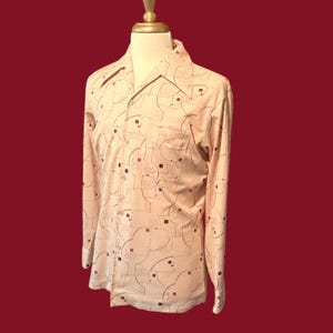 May include: A long-sleeved button-down shirt in a cream color, featuring a repeating pattern of abstract shapes in black, red, and gray. The shirt has a classic collar and a single chest pocket. The shirt is displayed on a mannequin.