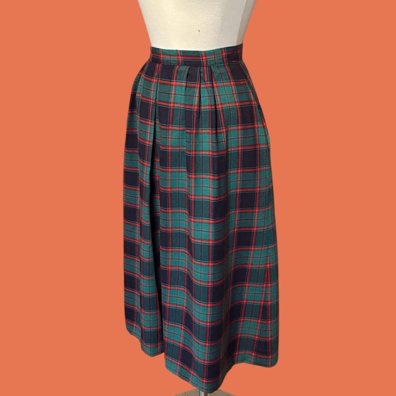 May include: A pleated, knee-length skirt in a classic plaid pattern. The skirt features a color scheme of teal, navy, and red. The garment is displayed on a mannequin against an orange background. The skirt is a vintage style.