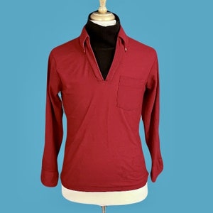 May include: A long-sleeved burgundy shirt with a v-neck collar and a single pocket on the left side. The shirt is worn over a black turtleneck.