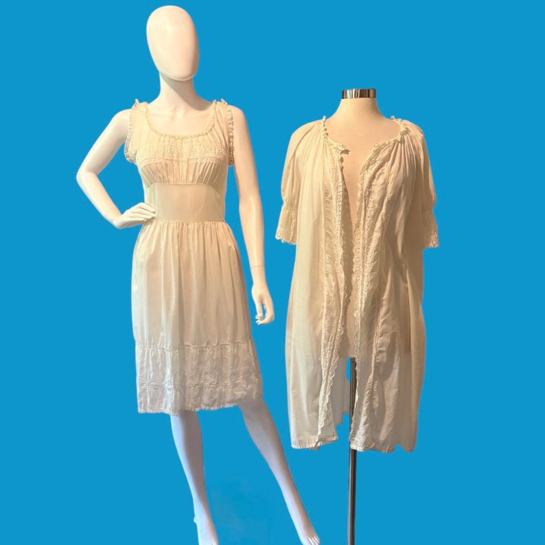 May include: Two vintage cream-colored dresses on mannequins against a blue background. One is a slip dress with a fitted bodice and a lace-trimmed hem. The other is a long, open-front robe with lace detailing.